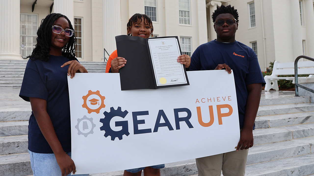 three students hold a banner for GEAR UP Achieve and show a formal document from the Alabama Senate declaring National GEAR UP Week while standing on the steps of the Alabama Capitol.