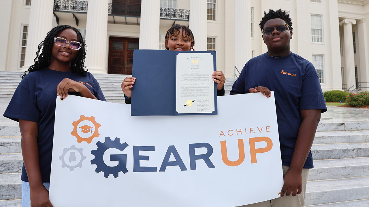 three students hold a banner for GEAR UP Achieve and show a formal document from the Alabama Senate declaring National GEAR UP Week while standing on the steps of the Alabama Capitol.