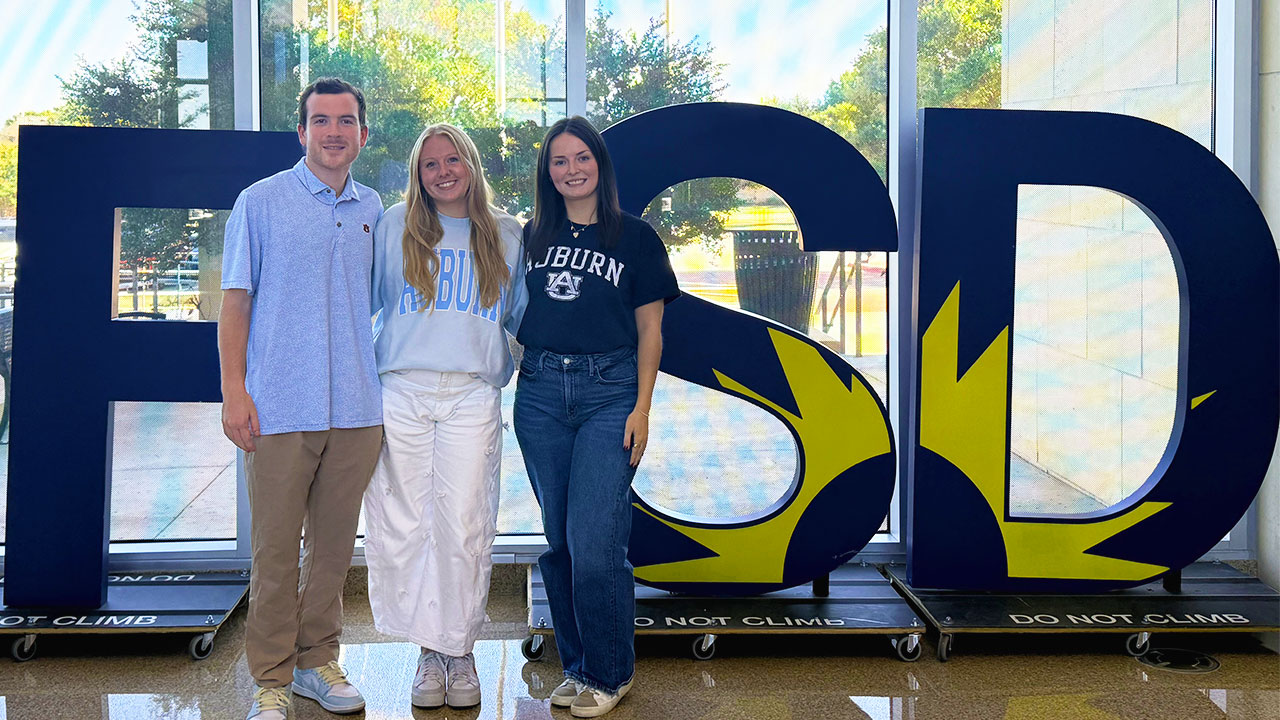 Three Auburn College of Education alumni stand in front of large letters representing the Frisco Independent School District for which they now work.