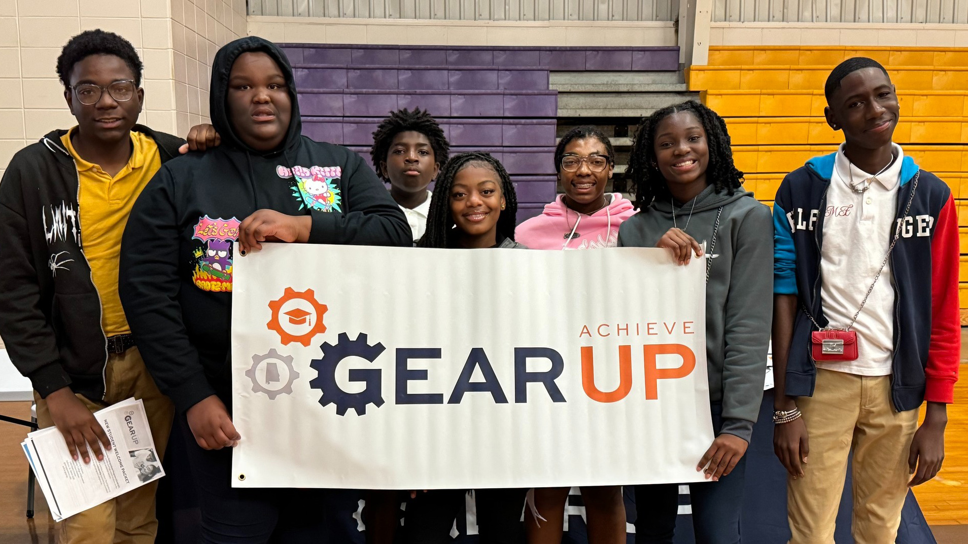 A group of GEAR UP Achieve students smile for a group picture while holding a GEAR UP Achieve logo banner.