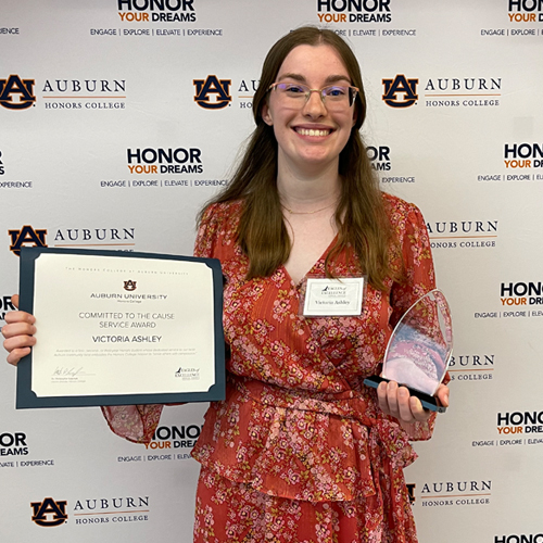 Victoria Ashley standing in front of the Honors step and repeat holding a certificate and an award