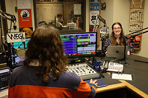 two girls a radio dj booth with radio equipment surrounding them there are colorful posters on the walls. One girl is speaking into the microphone and the other is working a display