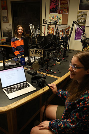 two girls a radio dj booth with radio equipment surrounding them there are colorful posters on the walls. One girl is interviewing a subject