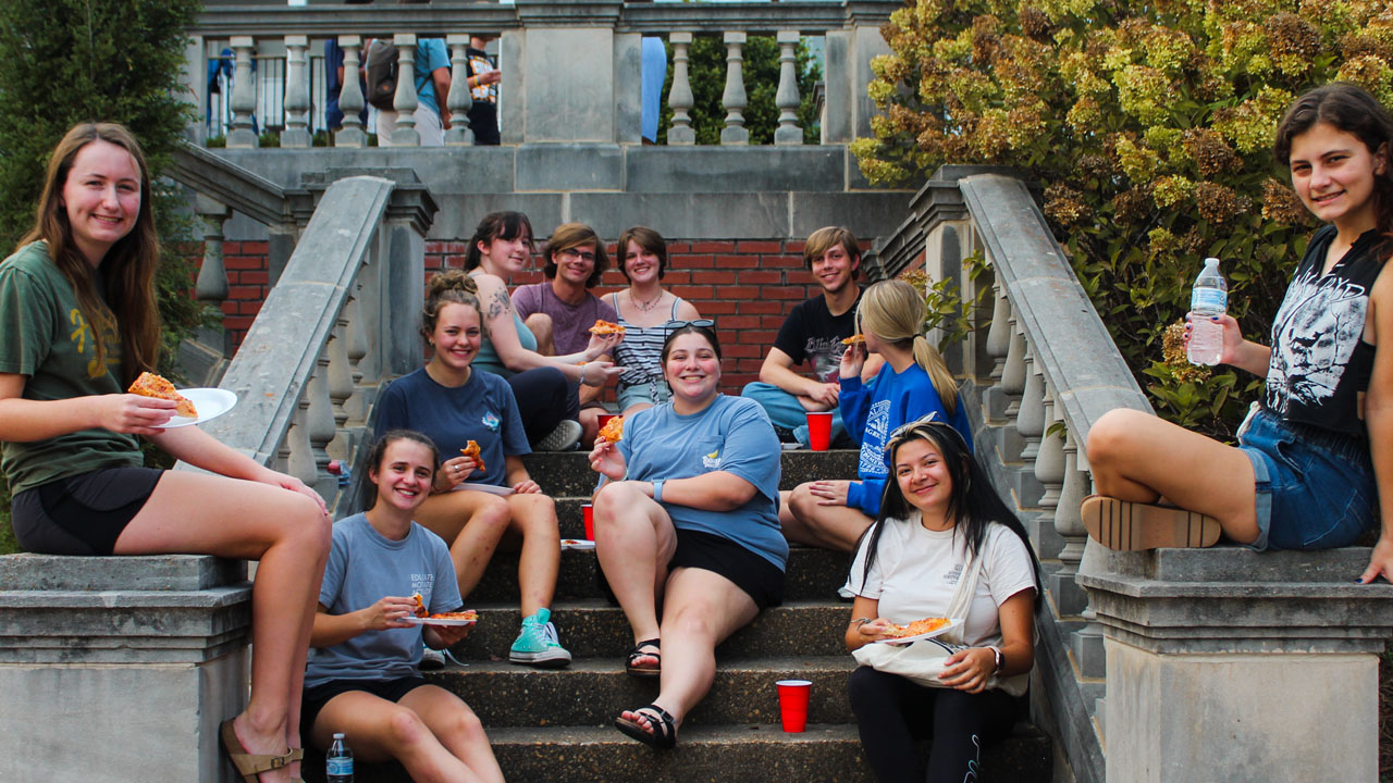 a group of students sitting on the back steps of Cater Hall enjoying food and drink