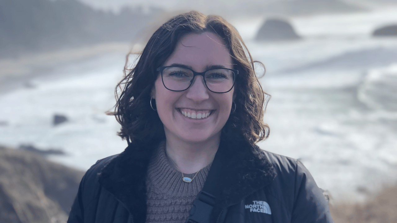 Headshot of Emily standing in front of a gray blue sky near the ocean. Her hair is blowing in the wind.