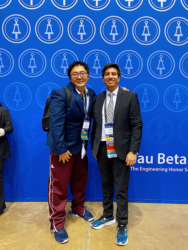 oliver at a conference in front of a blue background with another conference goer