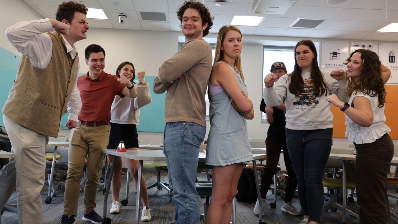 Eight students in a classroom. Two are standing in the center back to back with the other six are standing around them angrily.