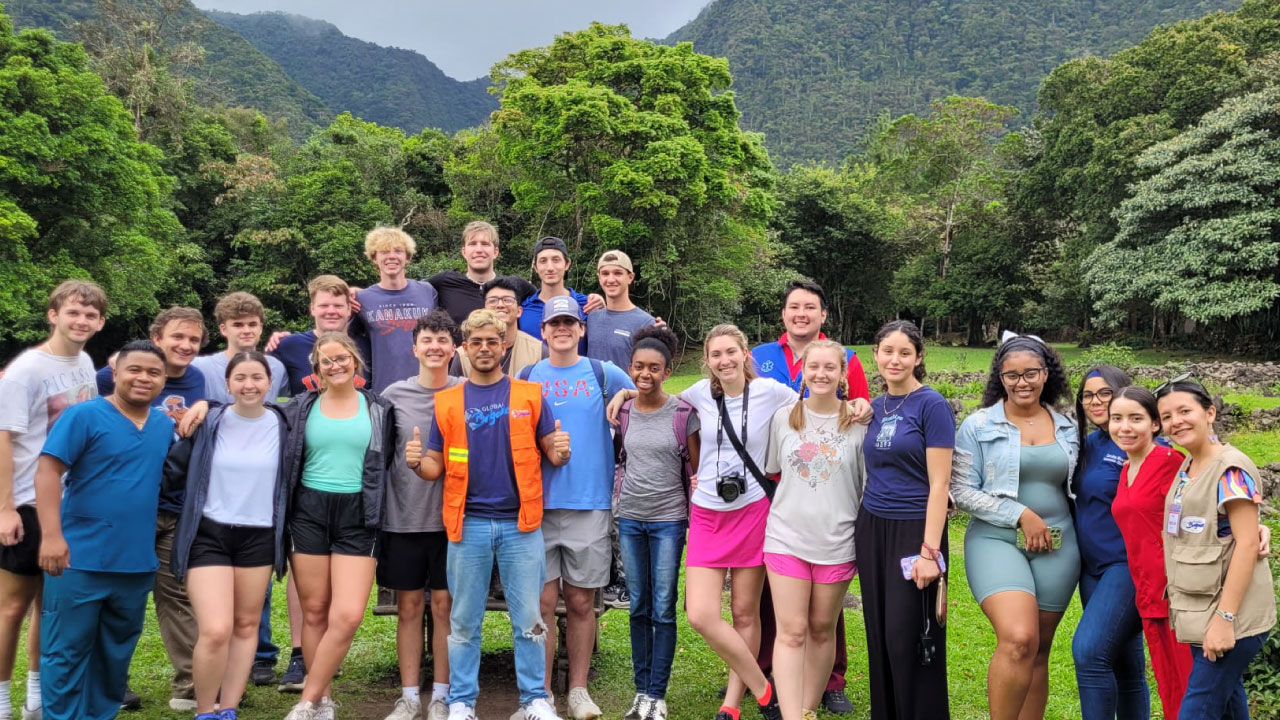 a large group of students standing in a tropical environment waving at the camera