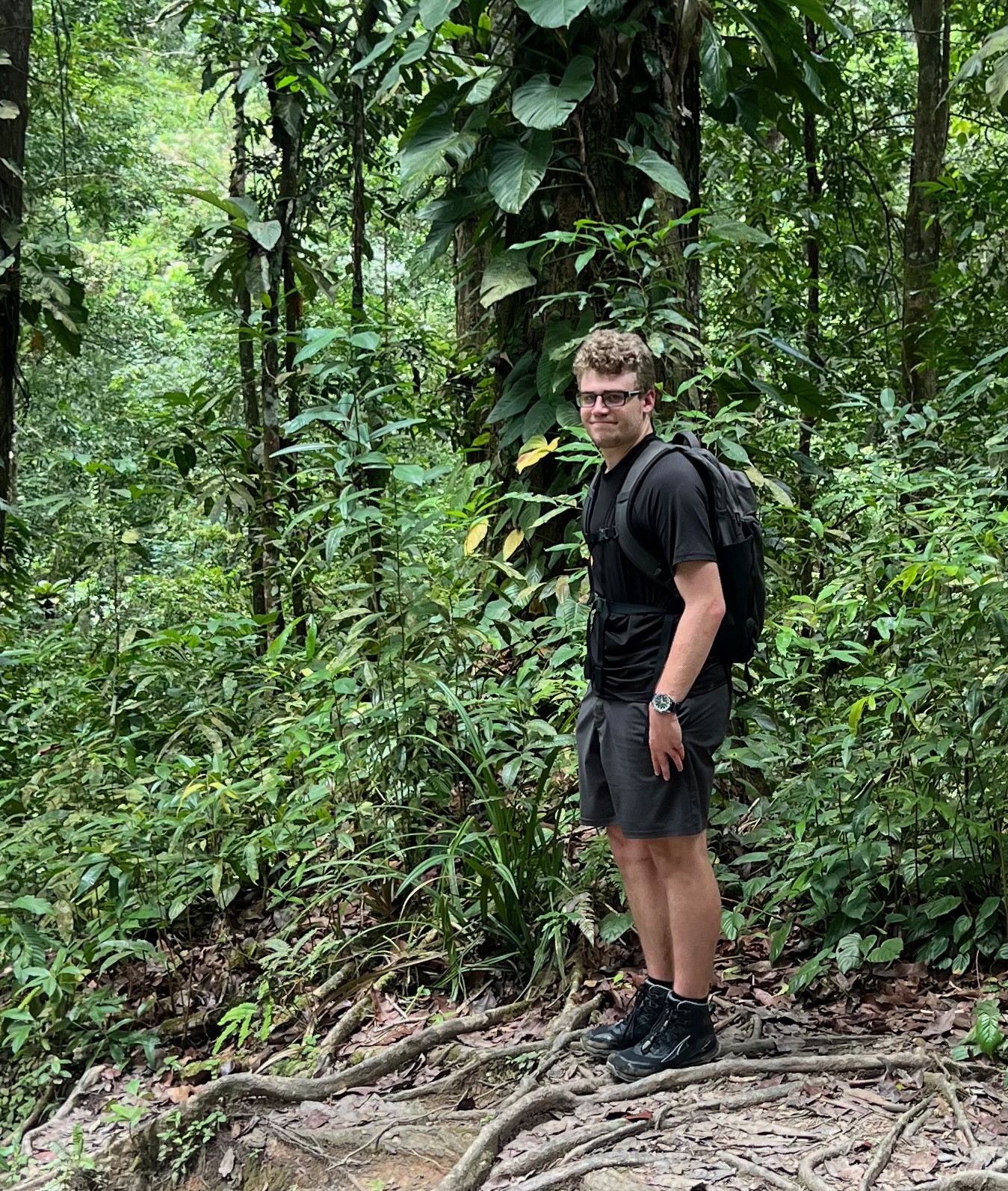 eirik is standing in a jungle with a black shirt and shorts on pointing at a tree