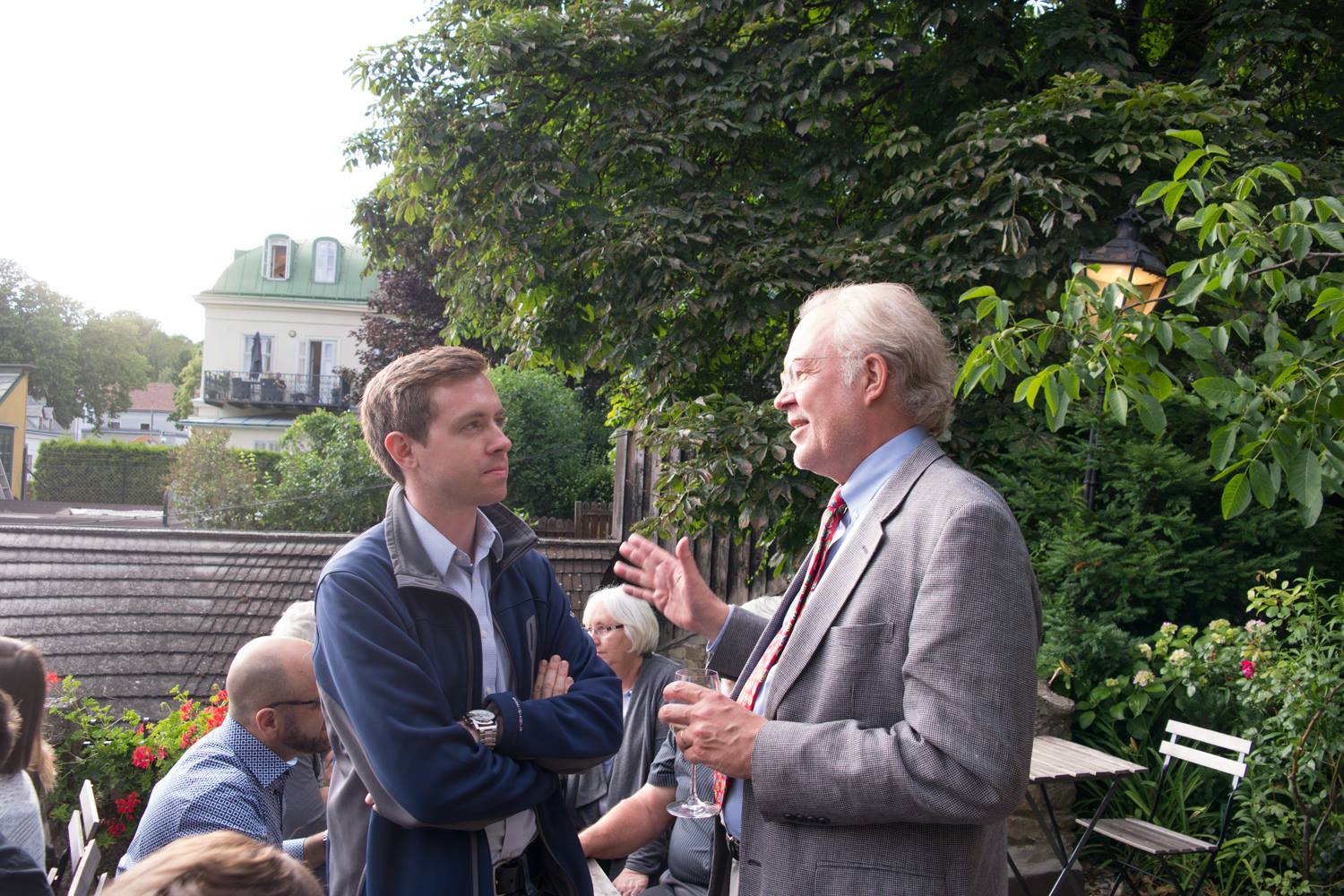 fagan is in a suit outside in a beautiful garden speaking with an older gentlemen in a tan suit