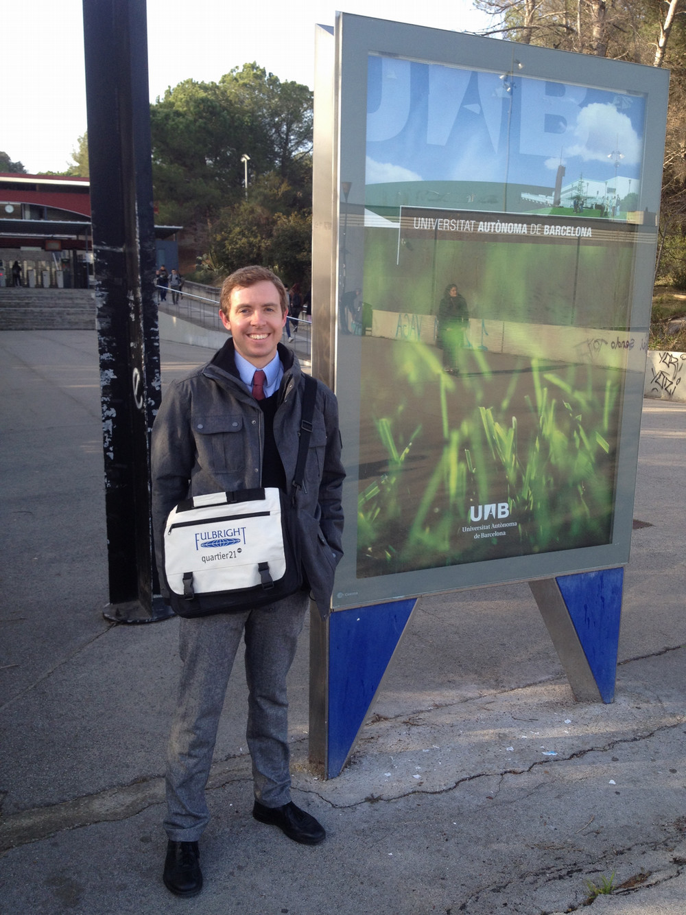 Ben is standing next to a sign in Barcelona in a blue jacket coat with a fulbright shoulder bag