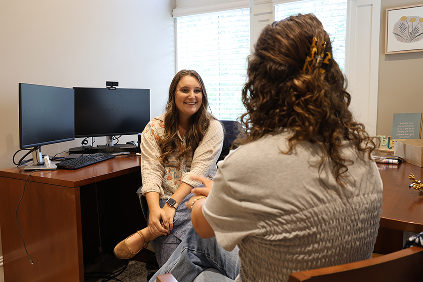 Two people are seated in an office, engaged in conversation. One person, facing the camera with a blurred face, wears a light-colored top and jeans. The other, with curly hair held by a clip and wearing a light gray top, has their back to the camera. A desk with dual monitors and various office supplies is visible in the background.