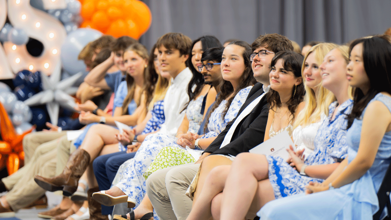 A group of individuals seated in a row at an indoor ceremony, dressed in semi-formal blue and white attire. Behind them are blue, silver, and orange balloons, along with large illuminated letters, creating a festive atmosphere