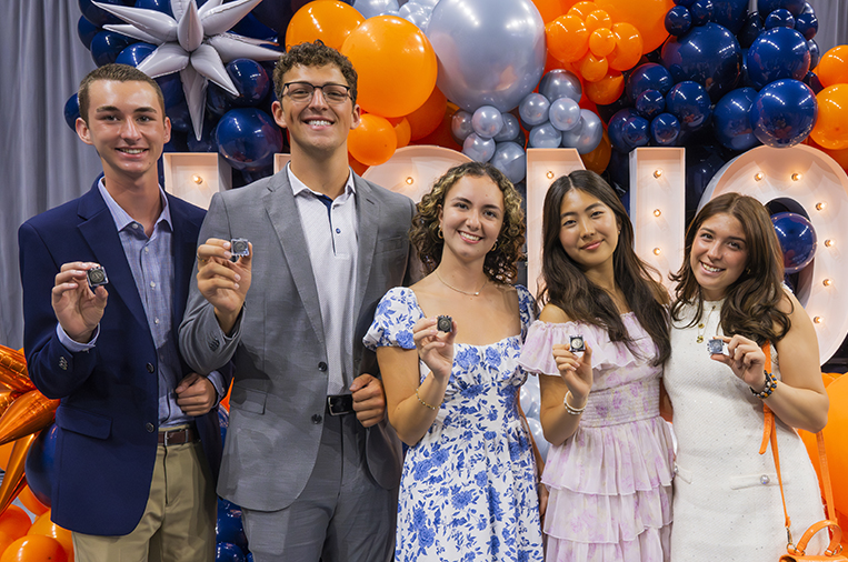 Five formally dressed individuals stand in front of a festive balloon backdrop featuring blue, orange, and silver balloons, along with large illuminated letters spelling 'HONORS.' Each person holds up an Honors pin.