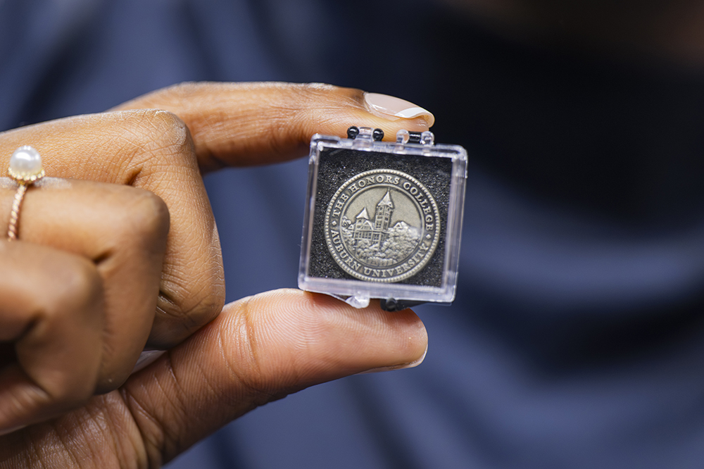 A hand holding a clear plastic case containing a round medallion. The medallion displays an embossed image of a building with the text 'THE HONORS COLLEGE OF AUBURN UNIVERSITY' around the edge, signifying academic recognition.