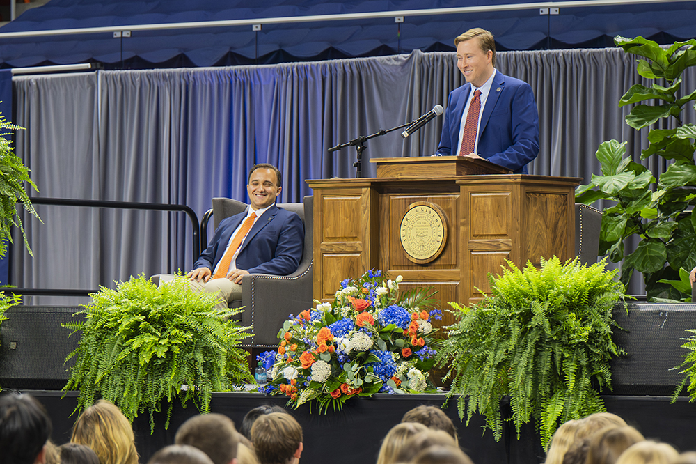 Two individuals on a stage during a formal event. One person is seated in an armchair on the left, while the other stands at a wooden podium with a microphone and emblem. The stage is decorated with green plants and a large floral arrangement of blue, white, and orange flowers. Grey curtains and blue seating areas form the background, with an audience visible in the foreground