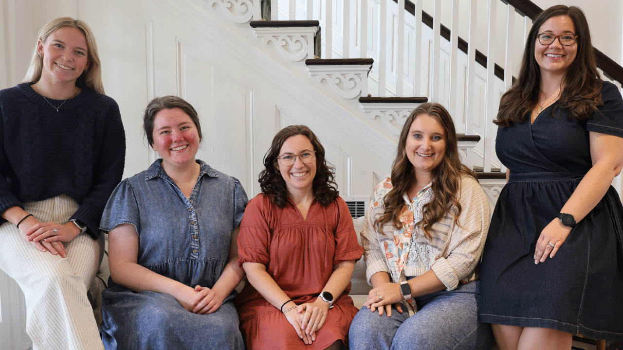 five women sitting on a couch. two are on the couch arms and three are sitting all are wearing causual clothing there is a wooden staircase behind them.