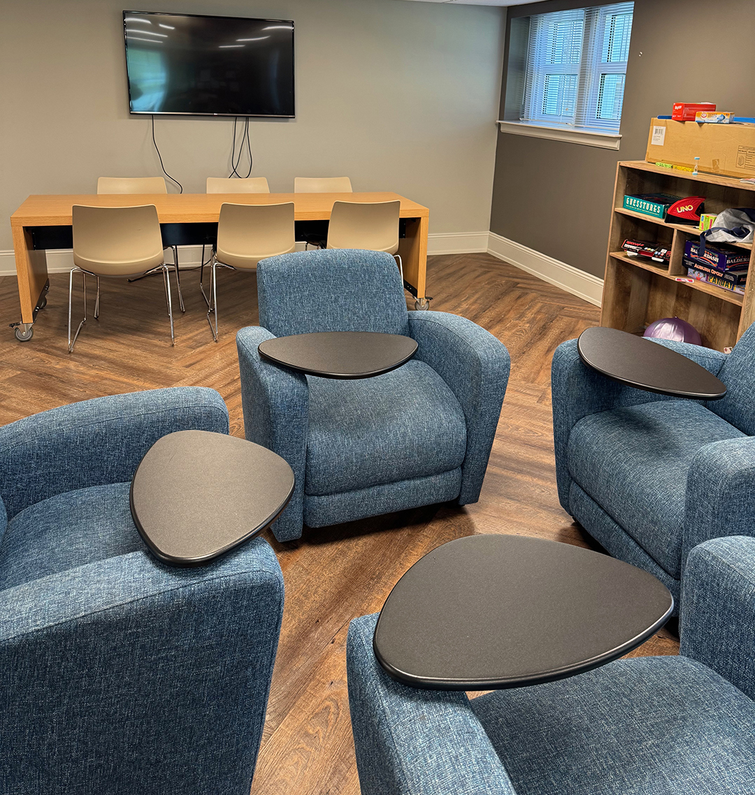 Room with four blue armchairs, each featuring a small black table attached to the armrest. In the background, a wooden table with four beige chairs sits beneath a wall-mounted flat-screen TV. A bookshelf on the right holds board games and other items. The room has wooden flooring and windows along one side.