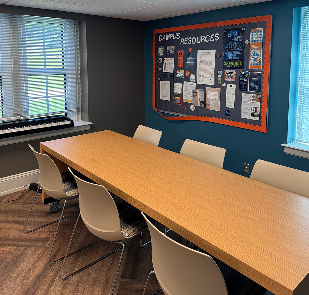 Conference room with a long wooden table surrounded by eight white chairs. A window with partially open blinds lets in natural light on the left, beneath which sits an electronic keyboard on a stand. On the right wall, a large bulletin board labeled 'CAMPUS RESOURCES' displays various flyers and informational materials. The room features wood-patterned flooring and dark gray and blue walls.