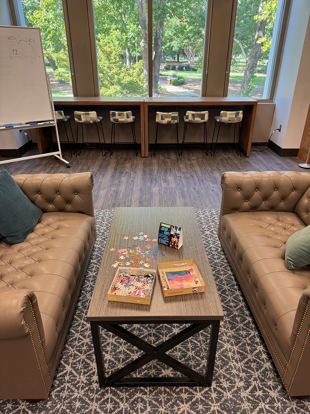Cozy room with two beige tufted sofas facing each other across a wooden coffee table holding two open puzzle boxes and scattered pieces. A large window in the background reveals green trees and a park-like view. Below the window, a long desk with stools is lined up. An easel whiteboard with drawings or writing stands to the left.