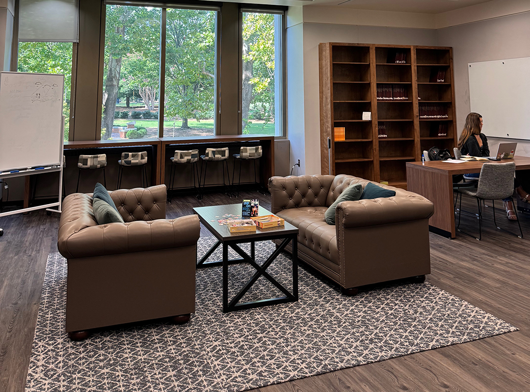 Cozy study or lounge area with two brown leather sofas facing each other across a coffee table holding books and snacks, set on a patterned rug. Large windows in the background reveal greenery outside. A whiteboard with faint writing stands to the left, and high stools line the window. A person works on a laptop at a desk near bookshelves and another whiteboard on the right