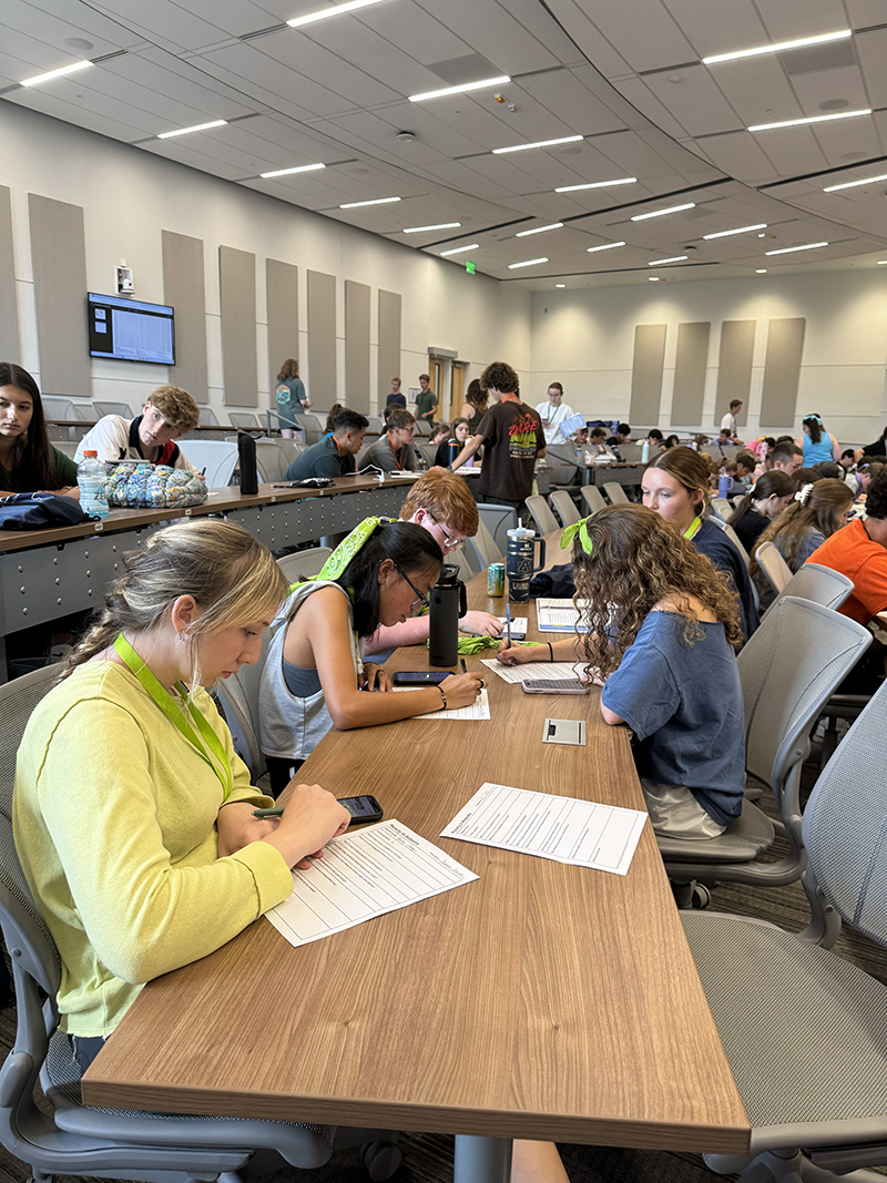 Classroom with students seated at long tables, working on assignments. Some have laptops and water bottles on the tables. The room features a modern design with a high ceiling, bright lighting, acoustic wall panels, and a large screen displaying information.