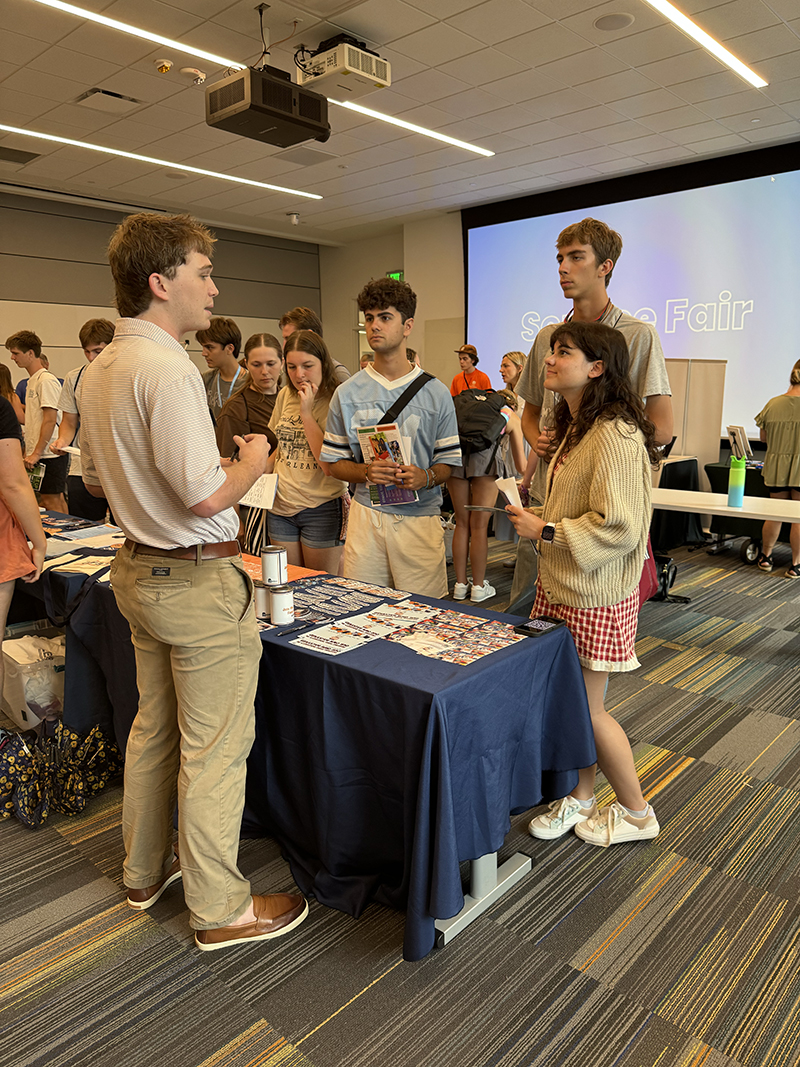 Group of people gathered around a table with a dark blue tablecloth at a school fair. The table displays pamphlets and materials, while a large screen in the background reads 'School Fair.' The room is brightly lit with overhead lighting, and other tables and attendees are visible in the background.