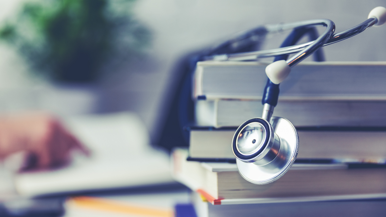 A stethoscope resting on a stack of books, with a blurred hand turning pages in the background, symbolizing medical education and healthcare studies.