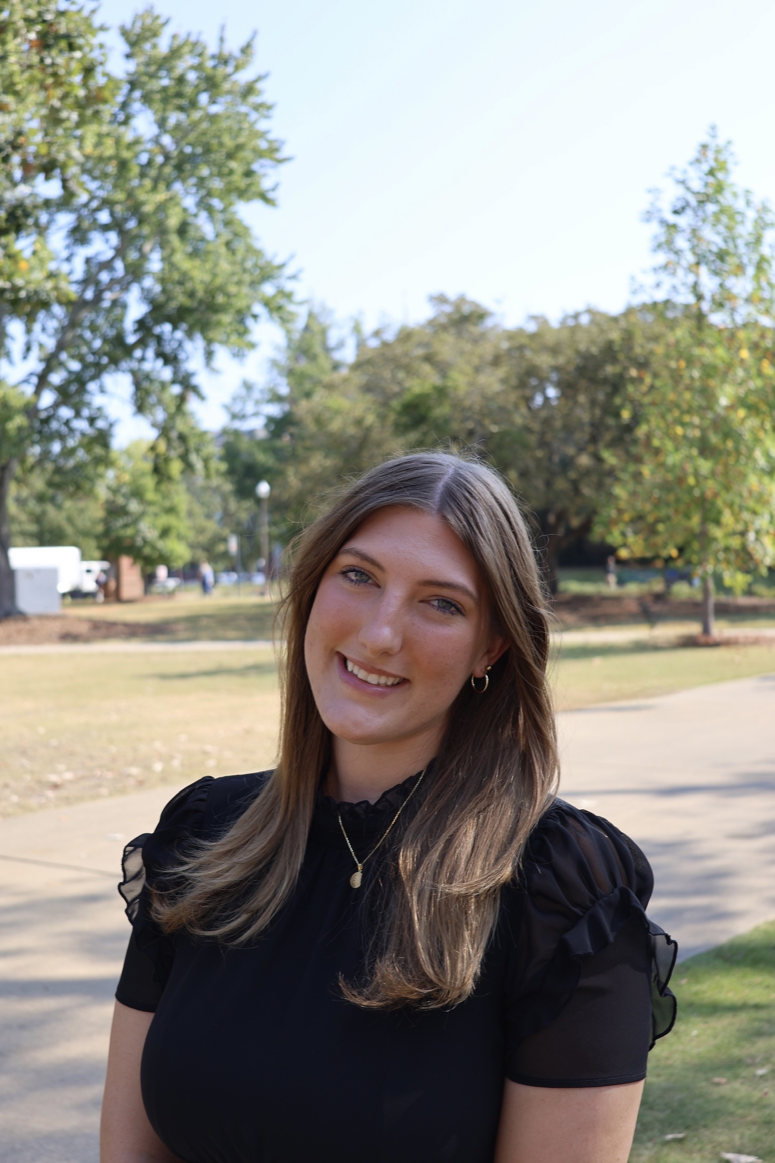 Person wearing a black short-sleeve top with sheer puffed sleeves and a gold necklace, standing outdoors on a paved path with green trees and grass in the background.