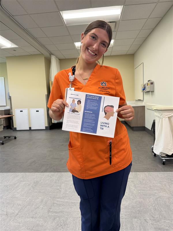 Person wearing an orange Auburn University nursing scrub top and navy blue pants, standing in a clinical setting and holding an informational pamphlet titled ‘Living with a TBI.