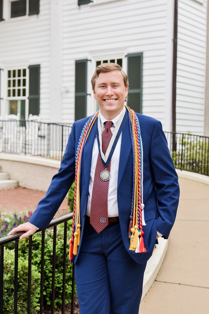 Jack is wearing a blue suit with a white dress shirt and a patterned red tie stands outdoors in front of a white building with green shutters. The individual has multiple colorful honor cords draped around the neck and a silver medallion hanging from a ribbon. One hand rests on a black metal railing along a curved walkway bordered by greenery.
