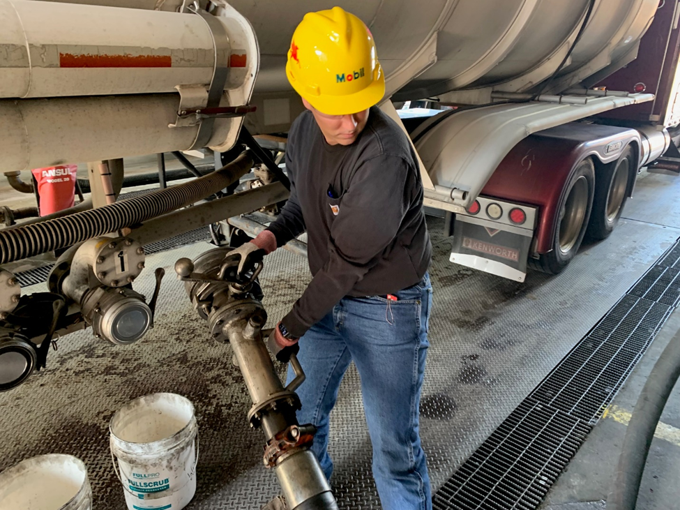 A worker wearing a yellow hard hat labeled “Mobil” and a dark long-sleeve shirt is connecting a large metal hose to a valve underneath a tanker truck. The scene appears to be in an industrial setting with a grated floor and safety equipment visible nearby. The tanker truck has multiple wheels and a cylindrical tank mounted on its chassis.
