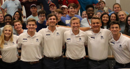 A group of people wearing matching white collared shirts with name tags and dark pants stand in a row with arms around each other in a classroom or meeting space. Behind them, more individuals are seated and standing, creating a crowded background.