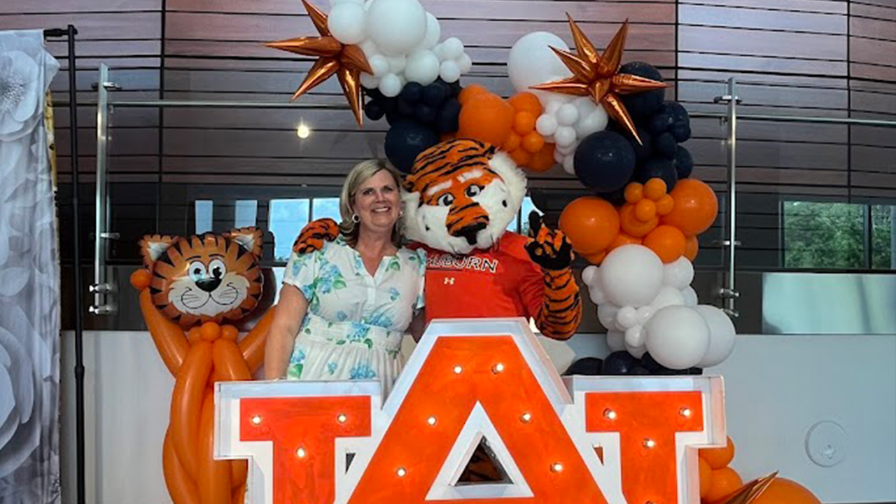 A blond woman wearing a blue and white dress poses with Aubie behind a big AU sign.
