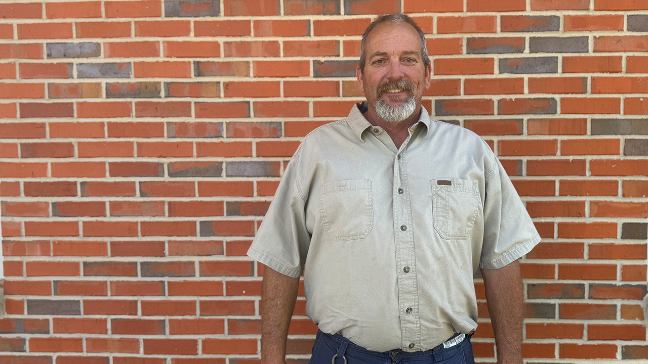 A gray haired man with a beard wearing a tan short sleeve shirt smiles for a photo while standing against a brick wall.