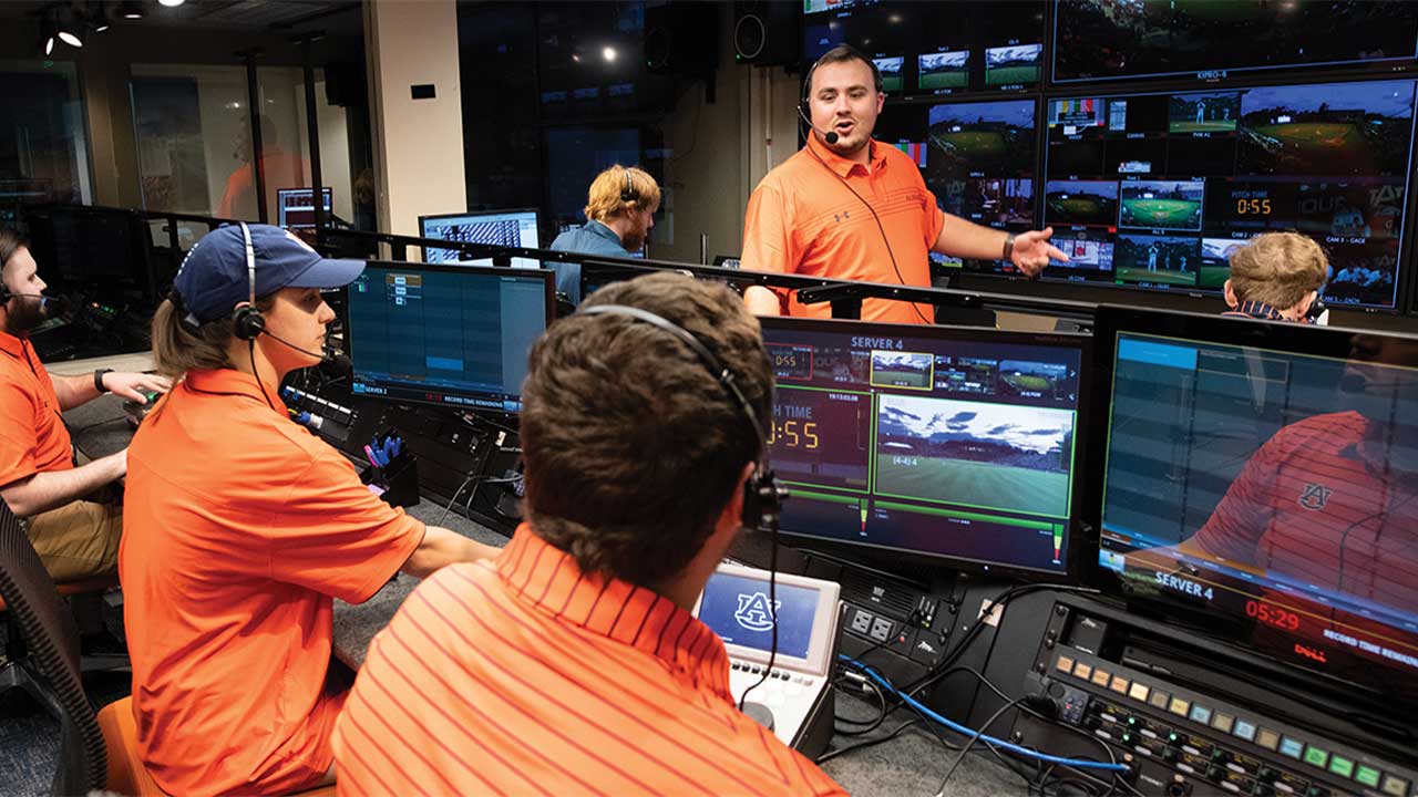 A group of people wearing orange shirts working in a broadcast control room with multiple screens displaying various camera feeds.