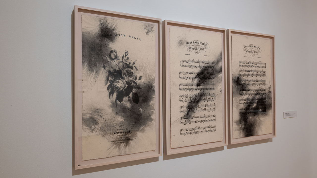 Framed sheet music, with charcoal markings, are displayed in a museum.