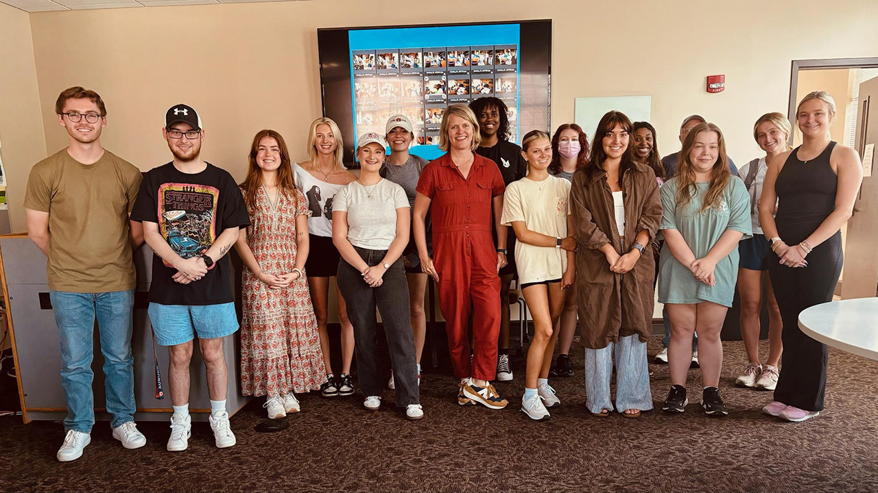Photo of AU undergraduate students in Advanced Reporting with photojournalist Kate Medley. Medley is in the center with short blonde hair and wearing a red jumpsuit.