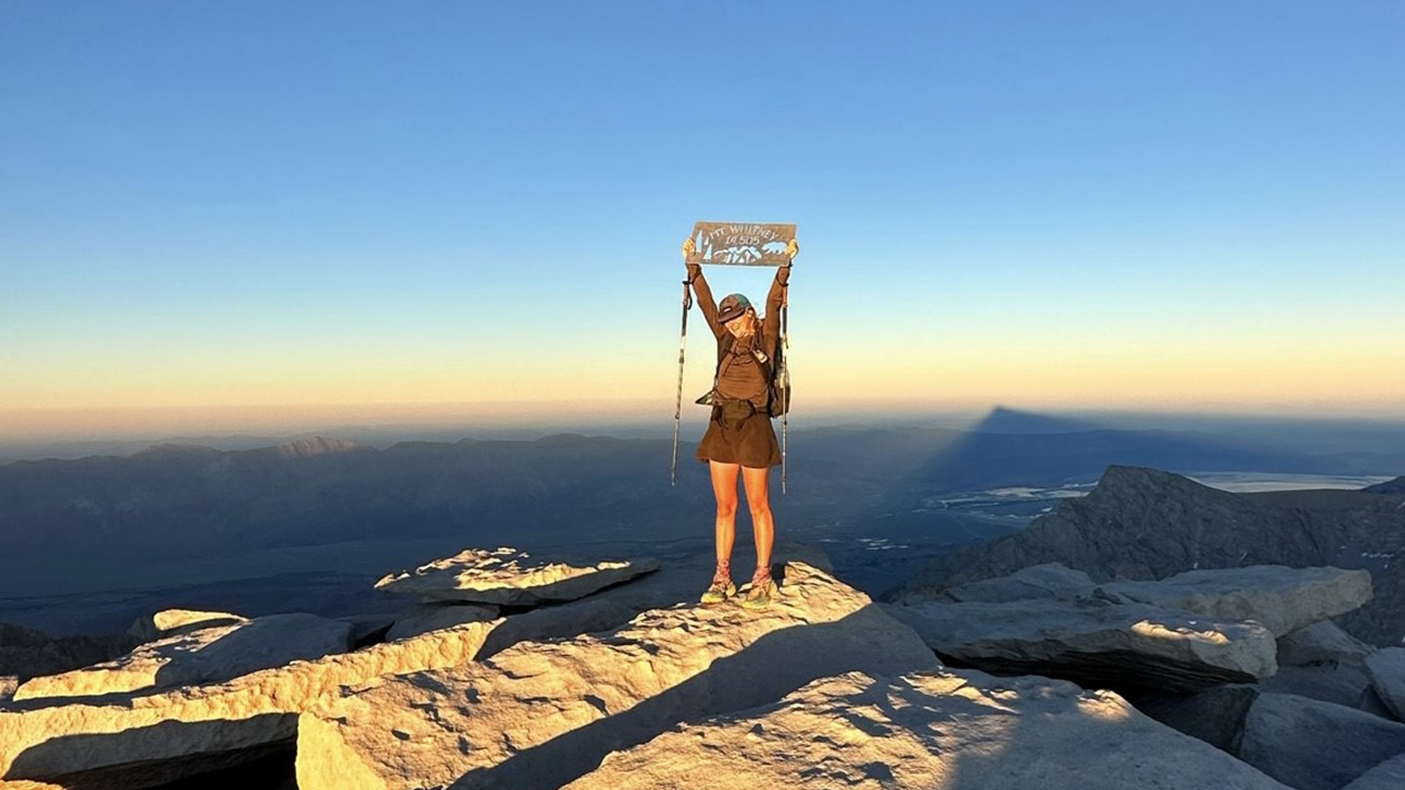 A young woman stands on top of a mountain with a colorful sunset in the sky behind her