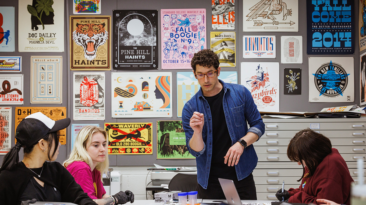 A man teaches a class of students with artwork hanging behind him