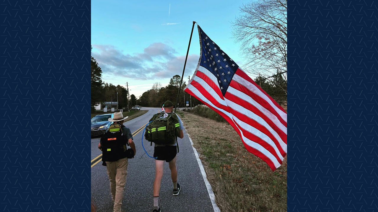 Two men walk on a road, one carrying the U.S. flag