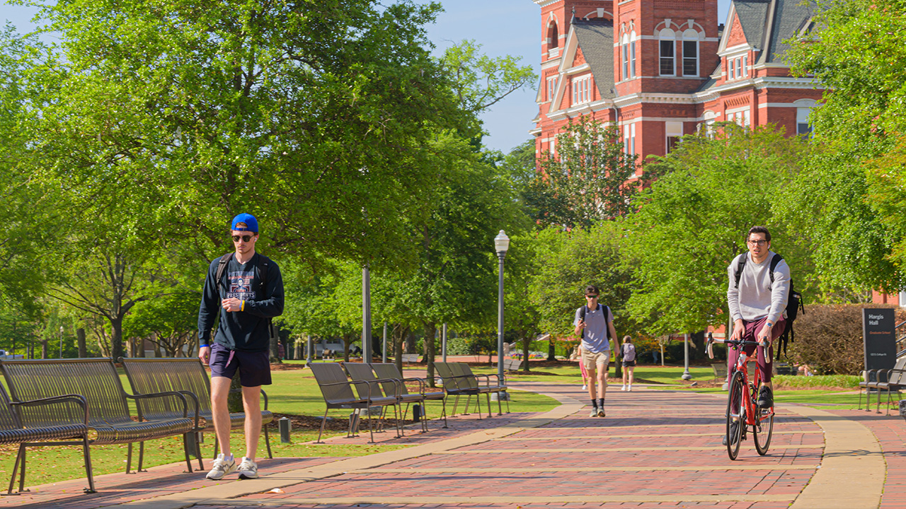 A person on a bike and several walkers travel on the sidewalk past Samford Hall