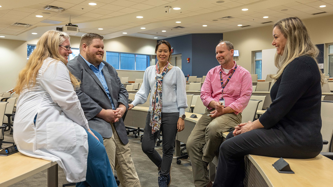 A group of five men and women sit on tables in a classroom talking and laughing