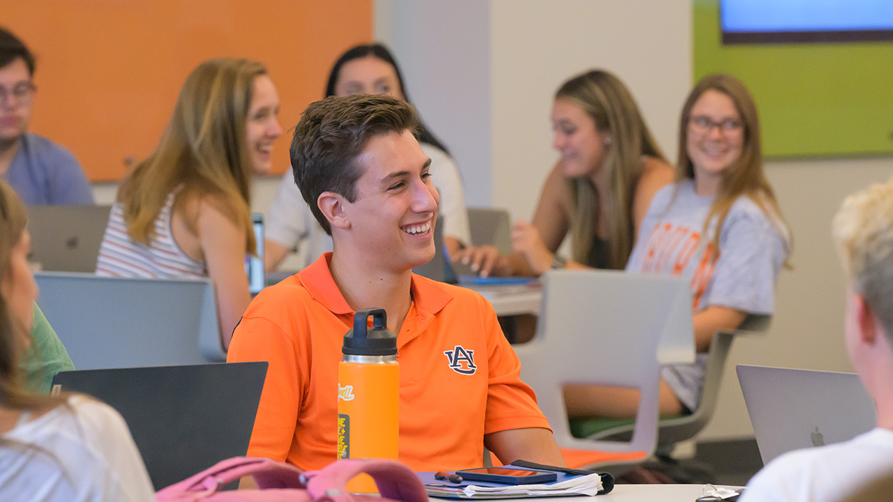 A young man in an orange Auburn shirt sits in a classroom laughing while other students around him laugh with him