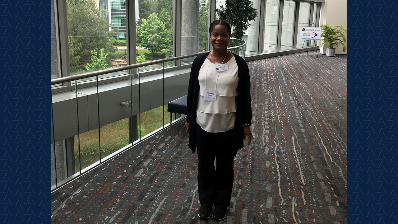 A young woman with professional clothing and a nametag stands in a hotel lobby smiling.