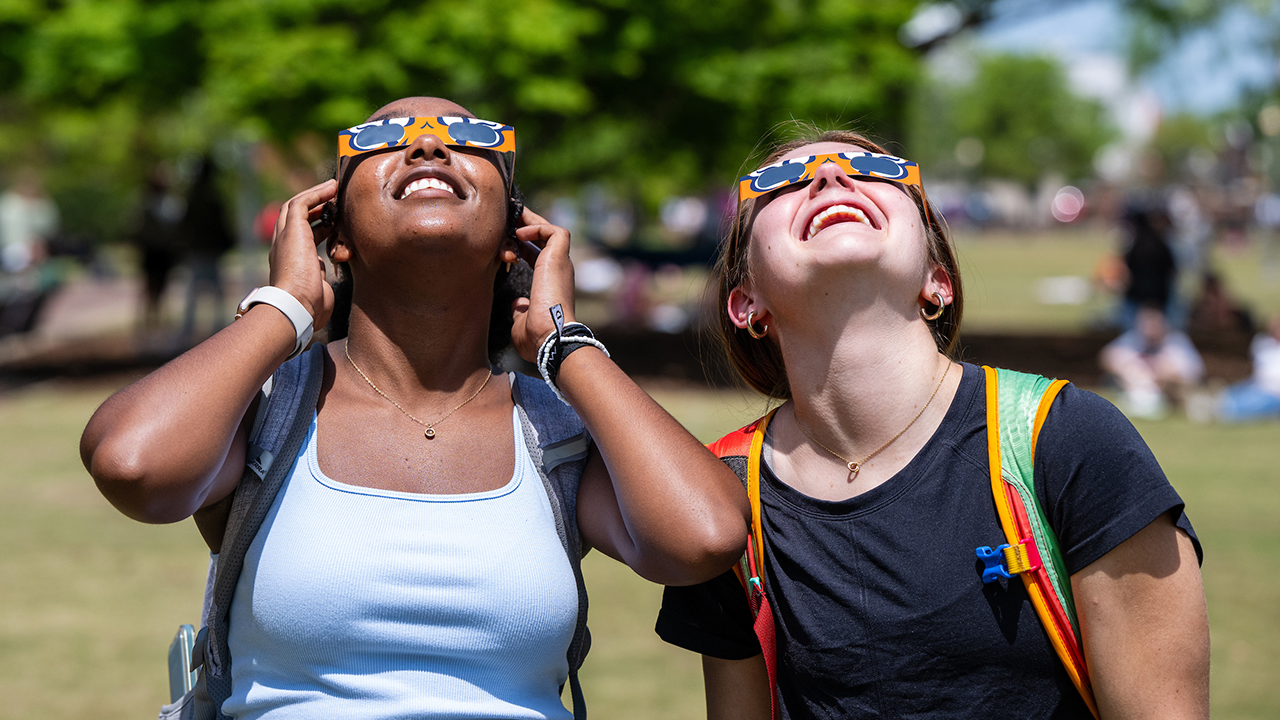Two students wearing eclipse glasses look up at the sun.