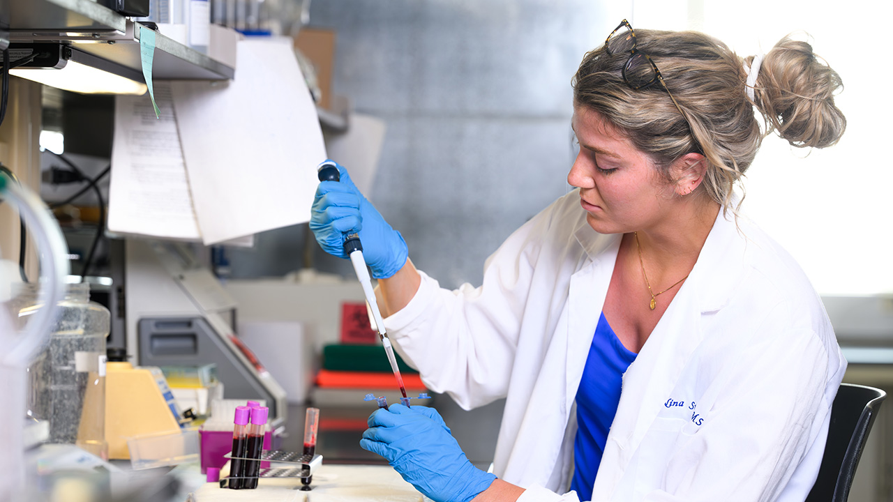 A woman in a lab coats uses a pipette to transfer fluid in to a test tube