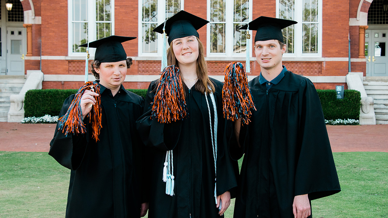 Three people in graduation caps and gowns wave orange and blue shakers