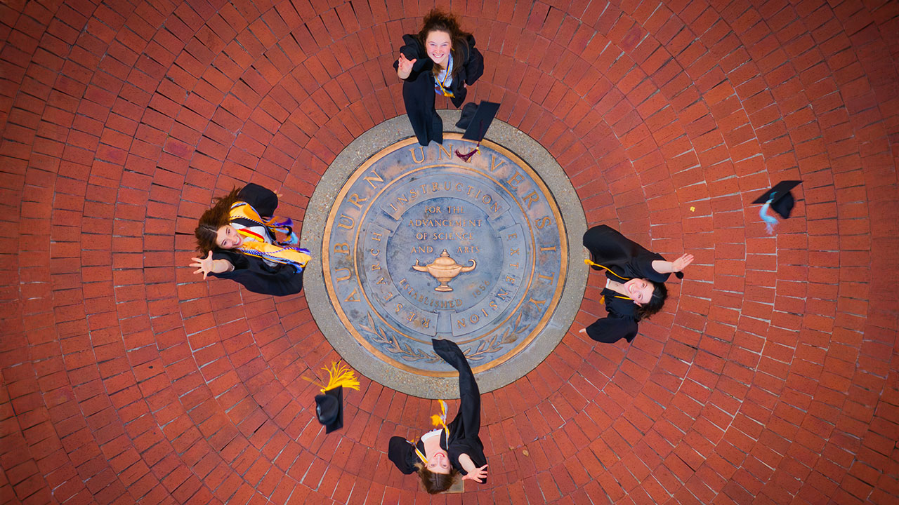 Four graduates through their caps in the sky at the Auburn University seal.