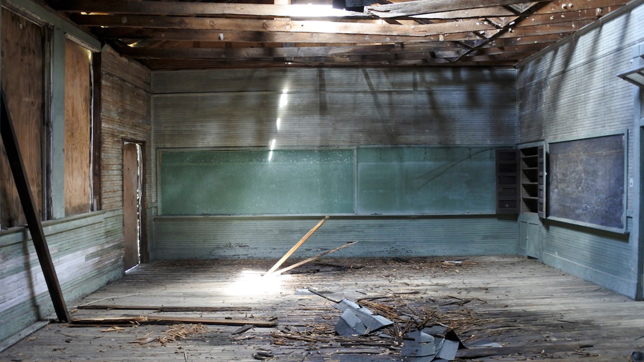 A bare room has a broken wood floor and damaged walls with a dim overhead light
